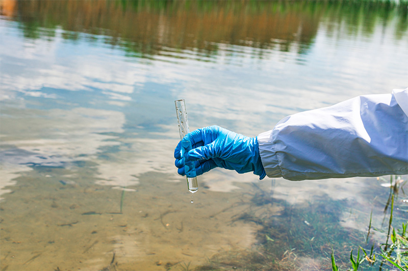 gloved hand holding a large test tube of water next to an outdoor water source